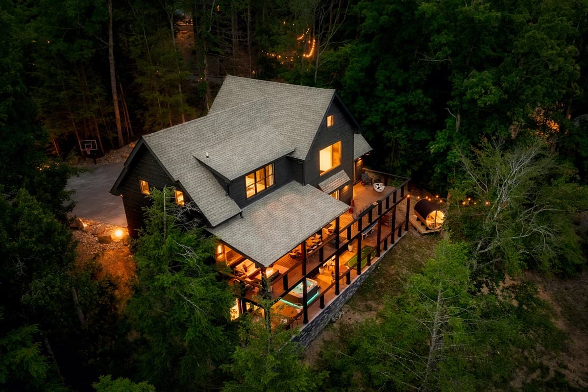 This luxurious home is nestled amidst a dense forest, featuring expansive decks and multiple levels, beautifully lit at night.