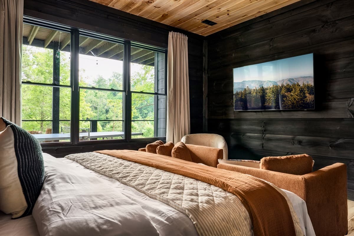 Relax and unwind in this beautifully designed bedroom, complete with a cozy bed featuring warm-toned bedding and a plush armchair, perfect for enjoying a book or the scenic outdoors. The room's sleek wall-mounted TV and large windows create a modern and se