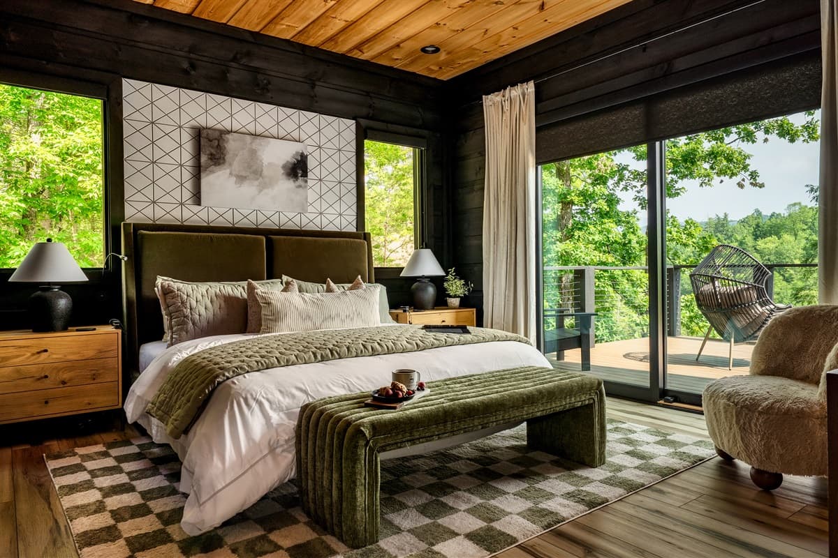 This stylish bedroom features a comfortable bed, warm wood ceiling, and large windows offering beautiful views of the surrounding trees.