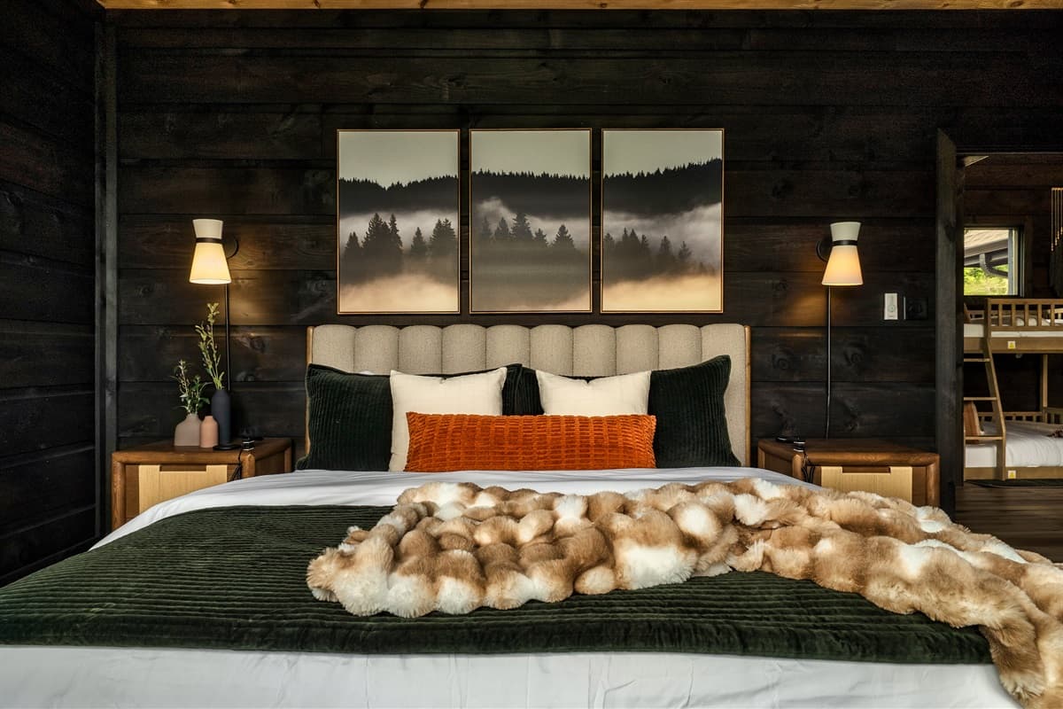 Relax in this beautifully appointed bedroom, complete with a large bed, stylish nightstands, and elegant wall sconces that illuminate scenic framed artwork above the headboard. The thoughtful design extends to the visible bunk room beyond, offering versati