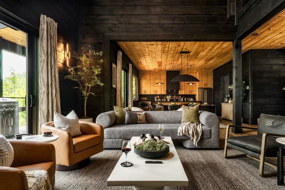 Relax and gather in this beautifully designed living room, offering a plush sofa, chic armchairs, and a warm ambiance created by the wooden ceilings and dark paneled walls. The open layout provides clear views into the adjacent well-appointed kitchen, maki