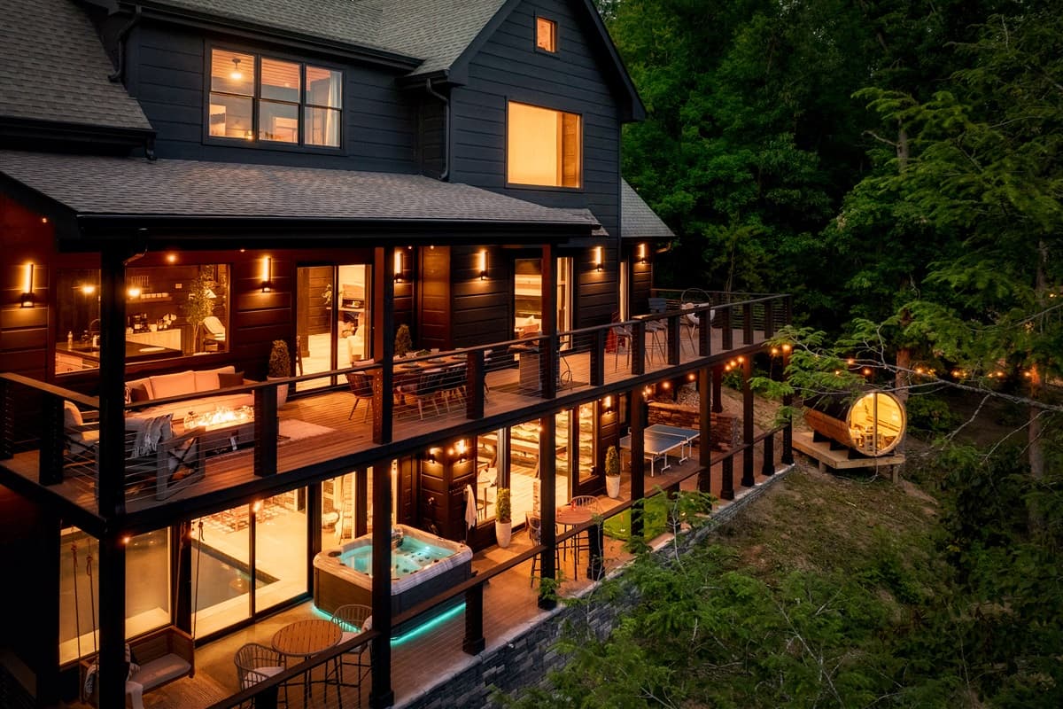 Experience luxury outdoor living on the spacious, illuminated decks of this stunning home, offering multiple areas for dining and lounging while surrounded by a serene wooded landscape. The warm glow of the exterior lights enhances the inviting ambiance fo