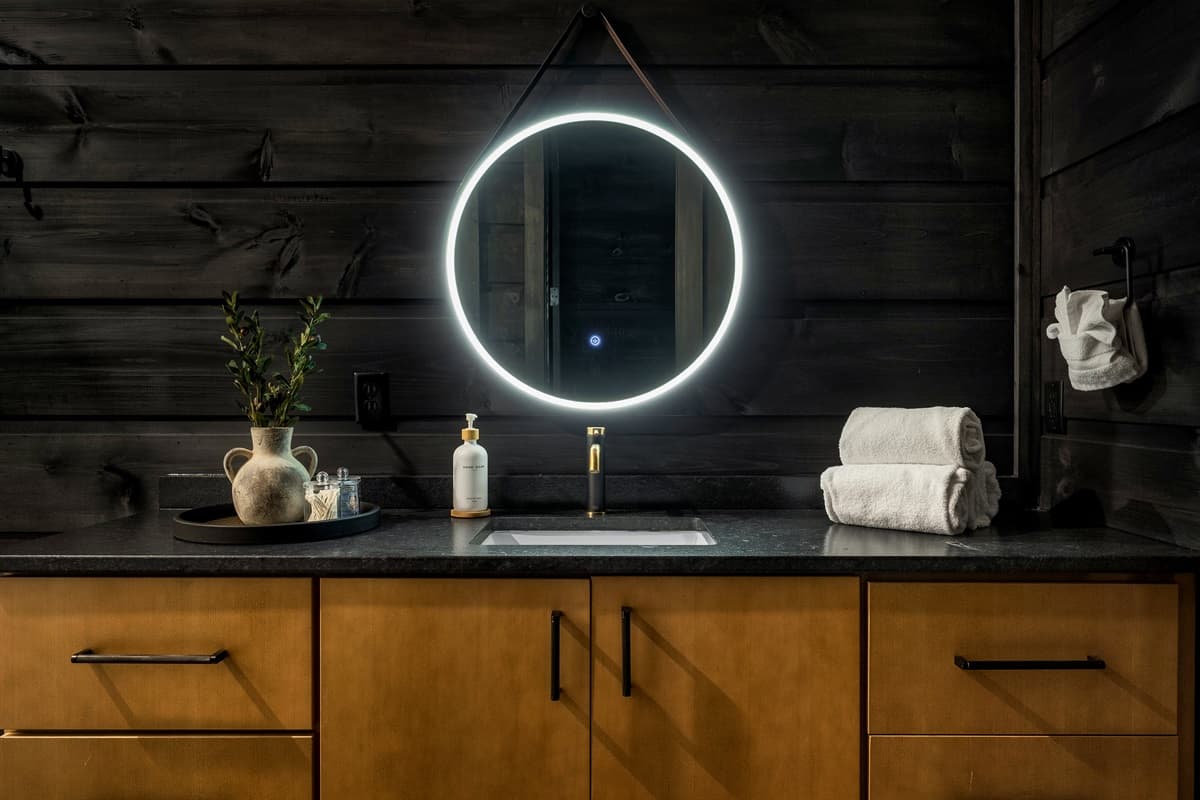 Experience contemporary comfort in this well-appointed bathroom, showcasing a minimalist sink set against dark wooden walls, brilliantly highlighted by a large circular mirror with integrated lighting. The thoughtful arrangement of soft towels and decorati