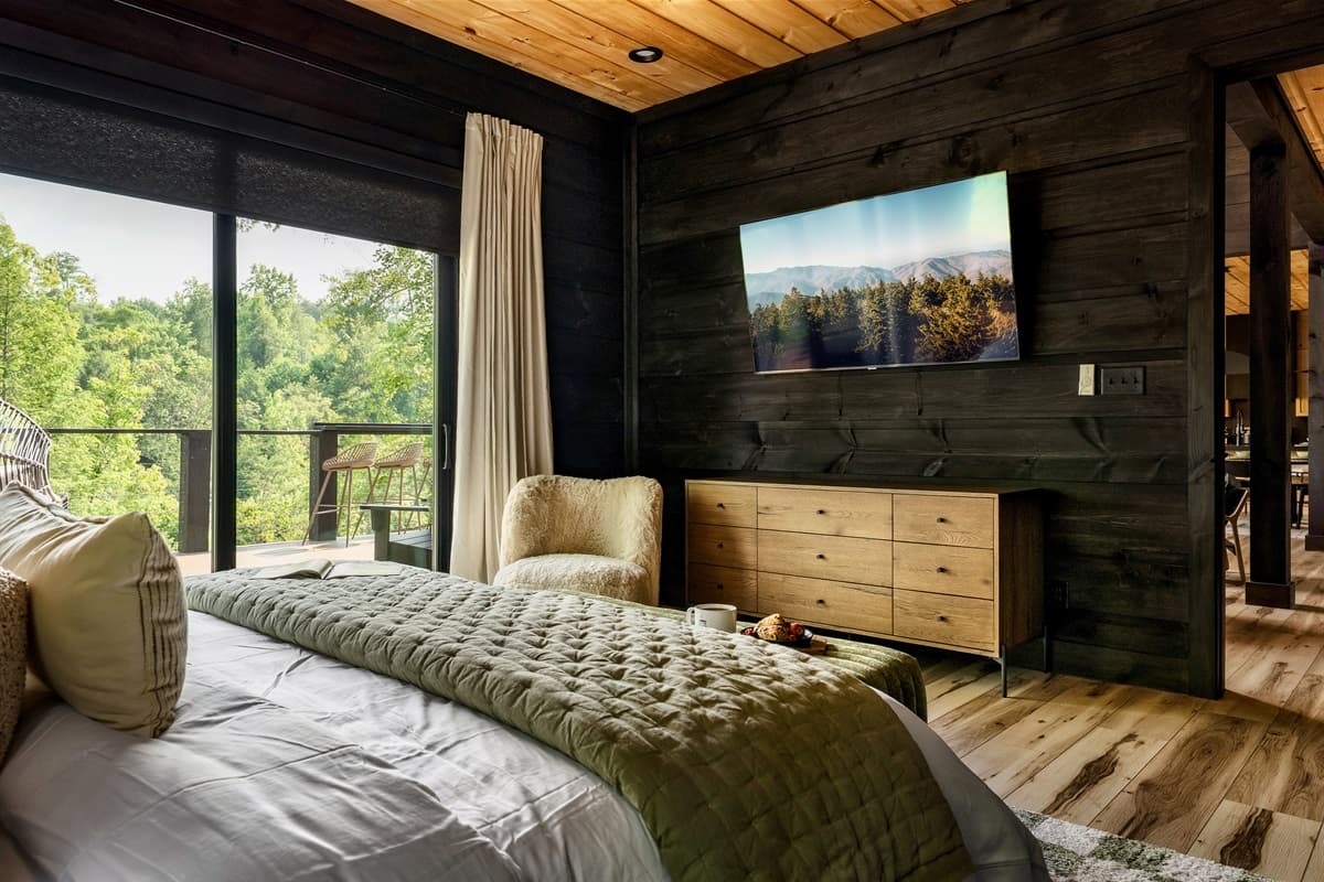 This spacious primary bedroom offers a comfortable bed, a wall-mounted TV for entertainment, and large windows with stunning views of the lush landscape.