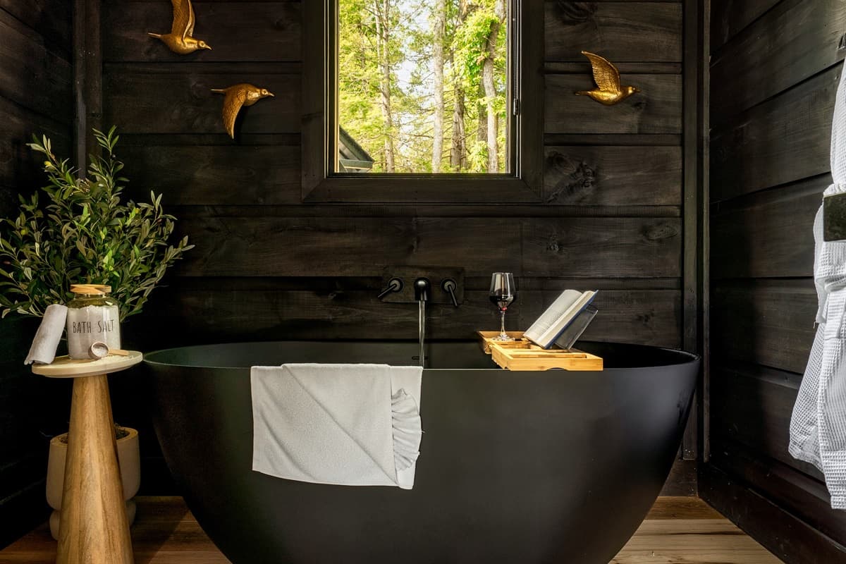This spa-like bathroom features a sophisticated black soaking tub, set against a backdrop of rich dark wooden walls, inviting ultimate tranquility. Thoughtful touches like a wooden bath caddy with reading material and a refreshing drink, alongside a verdan
