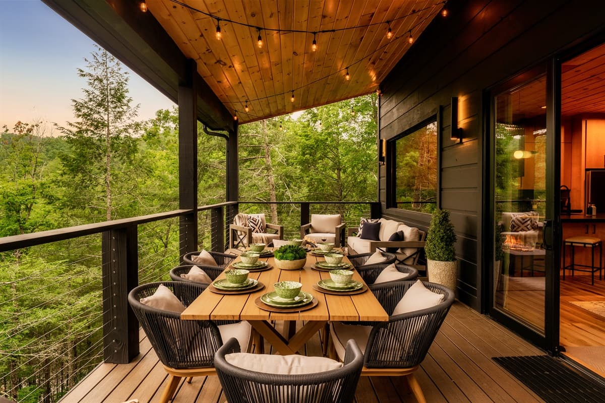 Enjoy al fresco dining surrounded by nature on this inviting outdoor deck, complete with a generously sized dining table, comfortable seating, and a warm ambiance from the overhead string lights. The beautiful treeline view provides a serene backdrop for a