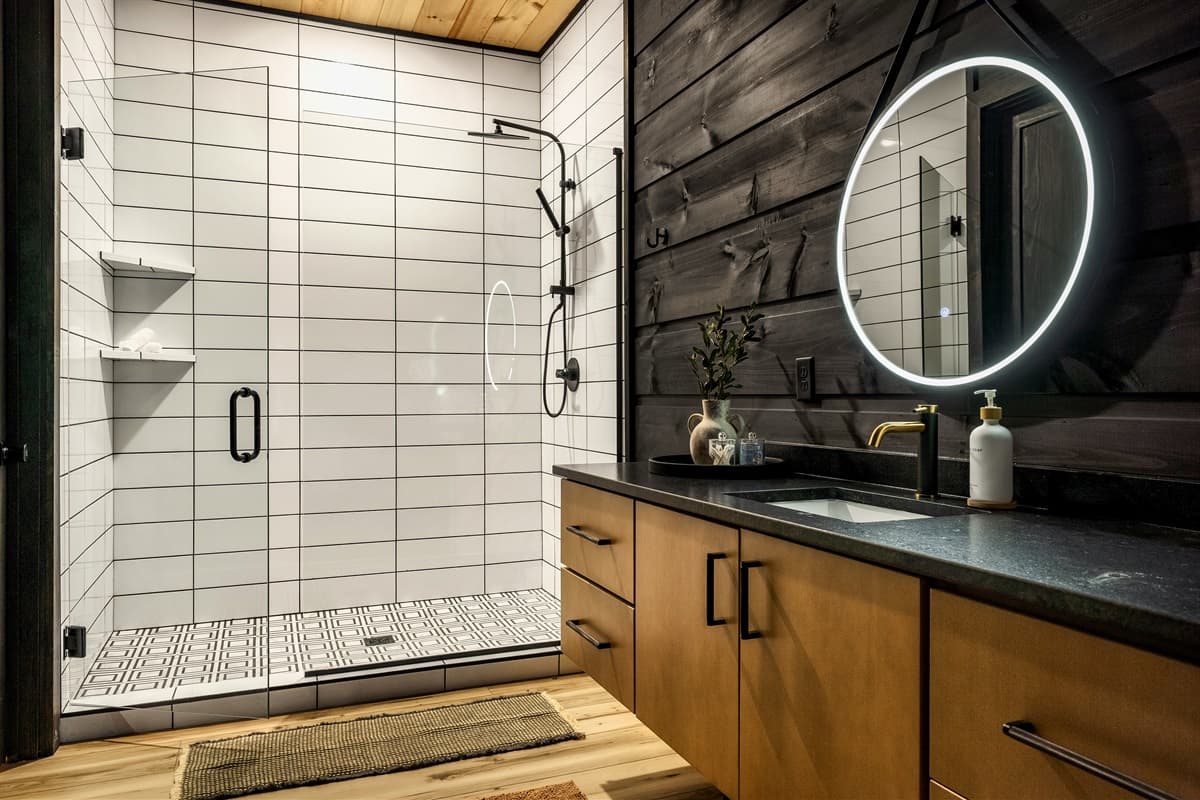 Enjoy a refreshing experience in this contemporary bathroom, which boasts a large walk-in shower with black fixtures and built-in shelves for convenience. The sleek wooden vanity with a dark countertop and a large, glowing circular mirror adds a touch of m