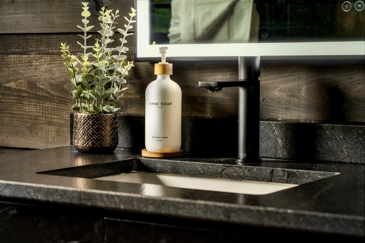 Modern convenience meets natural elegance in this bathroom, showcasing a dark countertop with a minimalist black faucet and a chic, eco-friendly soap dispenser. The lighted mirror and refreshing greenery add to the sophisticated and calming ambiance of the
