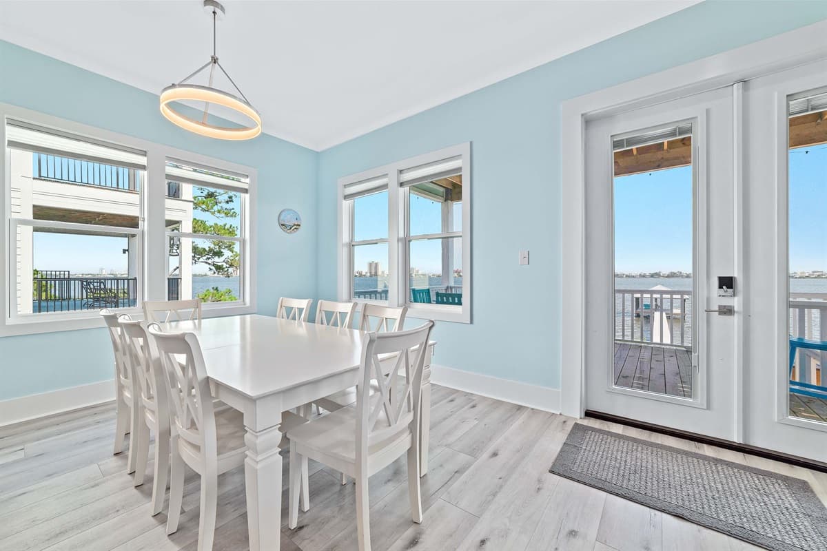 Enjoy bright, open-concept dining with a large white table and seating for eight, featuring views of the outdoors through multiple windows.