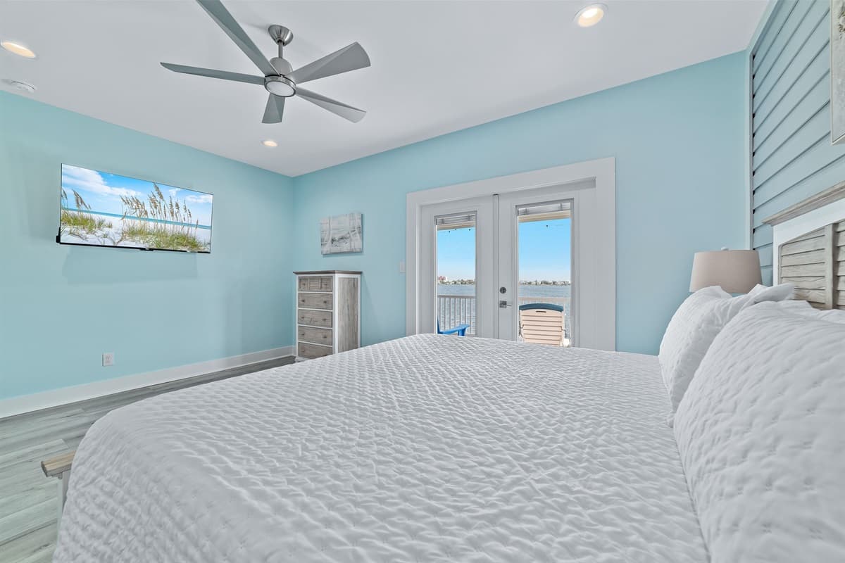 This bright bedroom features a comfortable king-sized bed, a wall-mounted TV, and direct access to an outdoor deck with water views.