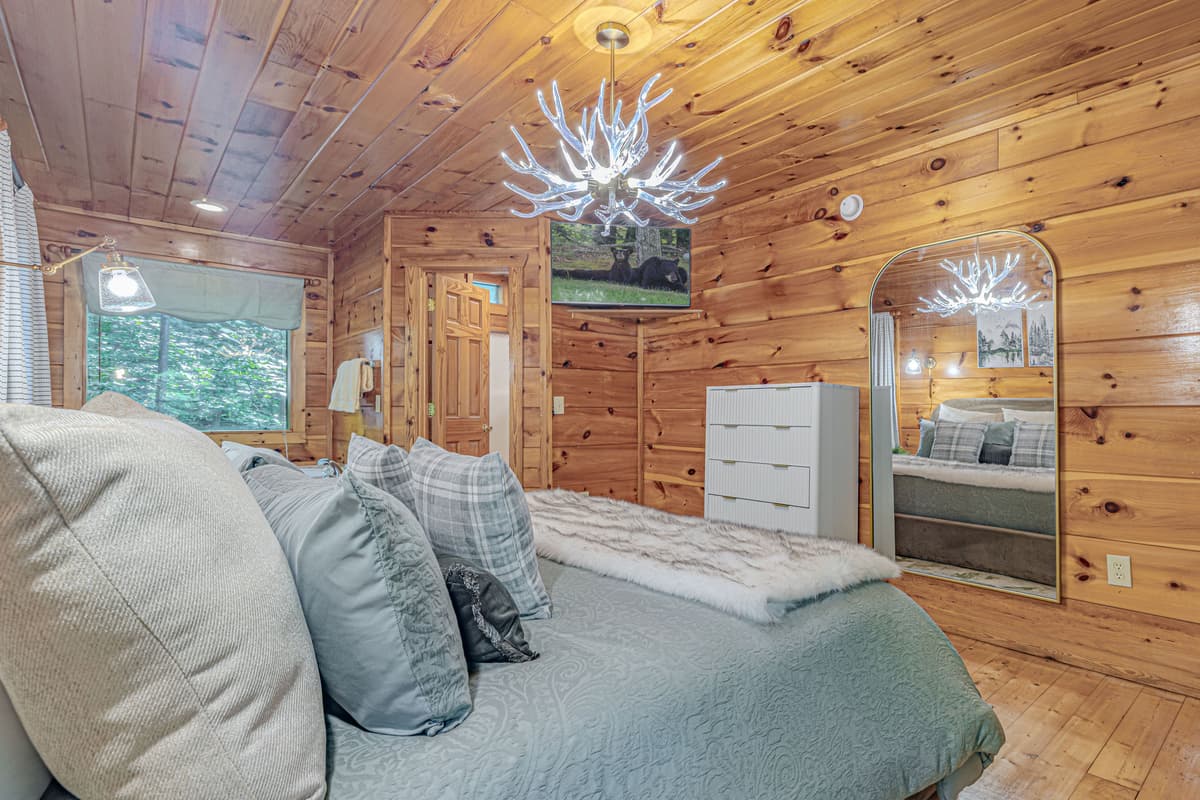 This inviting bedroom offers a serene escape, featuring a plush bed with soft linens, a large dresser for your belongings, and a mounted Smart TV for entertainment. The charming wood-paneled walls and rustic decor create a warm and cozy atmosphere.