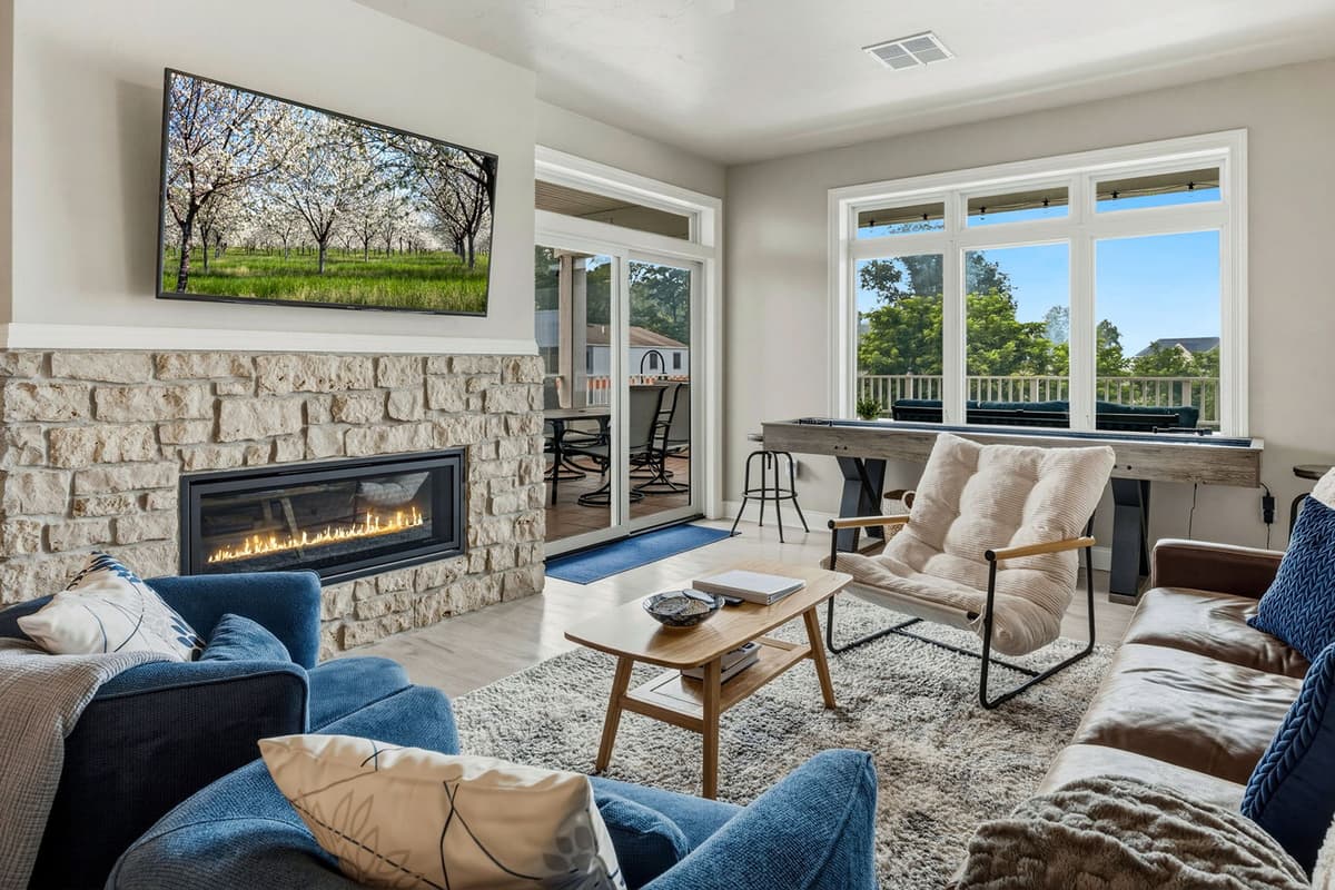 Relax in the spacious living area featuring comfortable seating, a TV, and a cozy fireplace. Large windows fill the room with natural light and offer beautiful views of the surrounding trees and greenery.