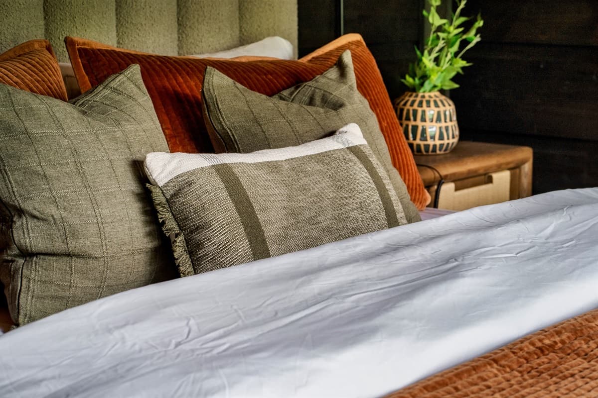 A close-up of a well-dressed bed reveals a comfortable mix of plush pillows in earthy tones and a decorative vase with greenery.