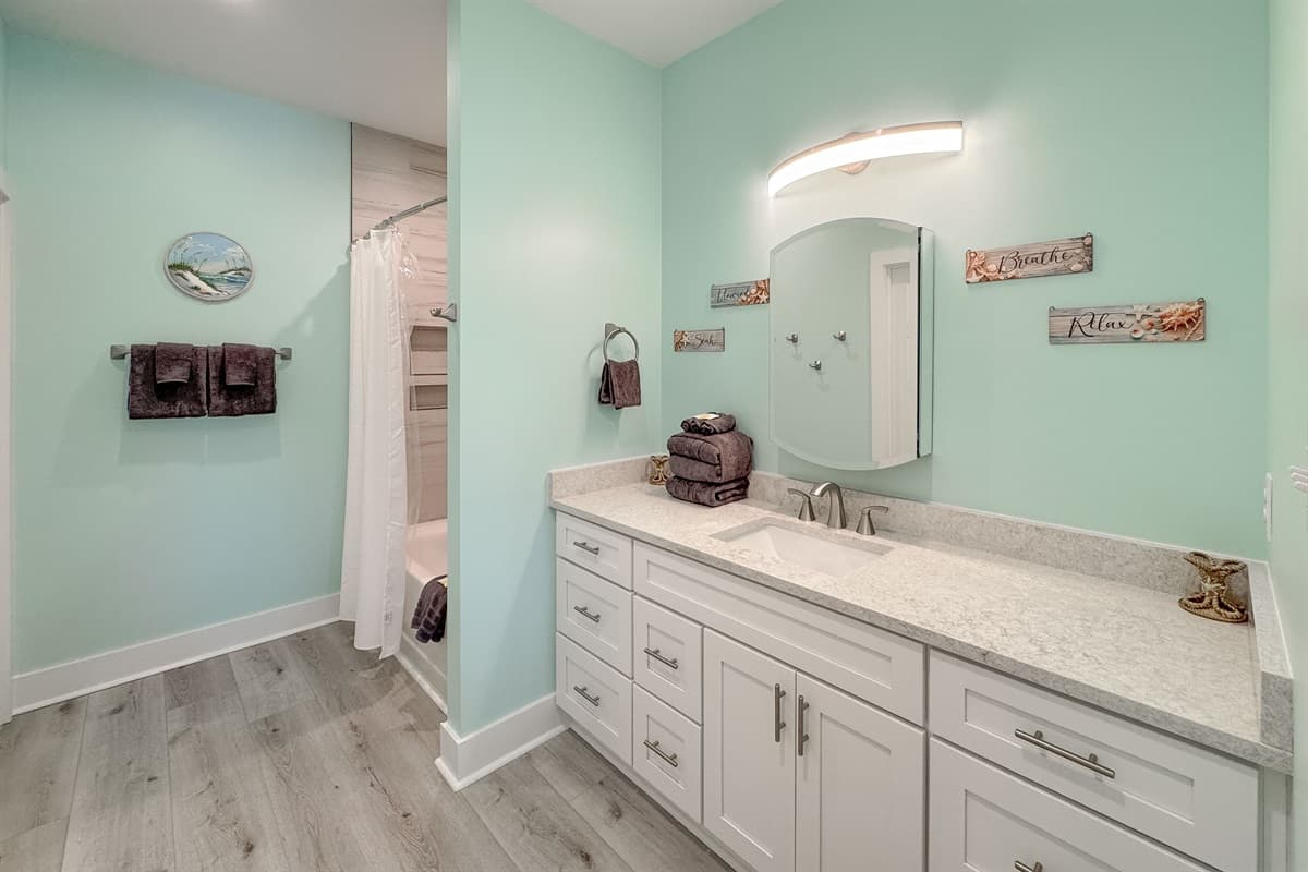 Enjoy the clean and inviting ambiance of this bathroom, offering a generous white vanity with ample counter space and soft lighting above a large framed mirror. The room includes a convenient combination tub and shower, with subtle coastal-themed decor on