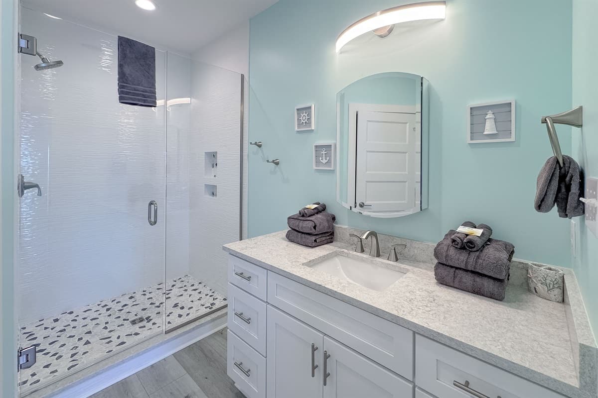 Step into this refreshing bathroom, designed with a contemporary vanity, elegant light green walls, and a large mirror. The highlight is the generous walk-in shower, featuring crisp white subway tiles and a distinctive pebble-style floor for a spa-like fee