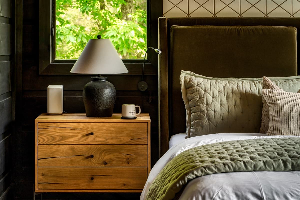 This inviting nightstand vignette offers both style and convenience, with a modern lamp providing soft illumination and a small mug ready for a relaxing drink. The warm wooden tones of the nightstand beautifully complement the dark paneled walls, creating
