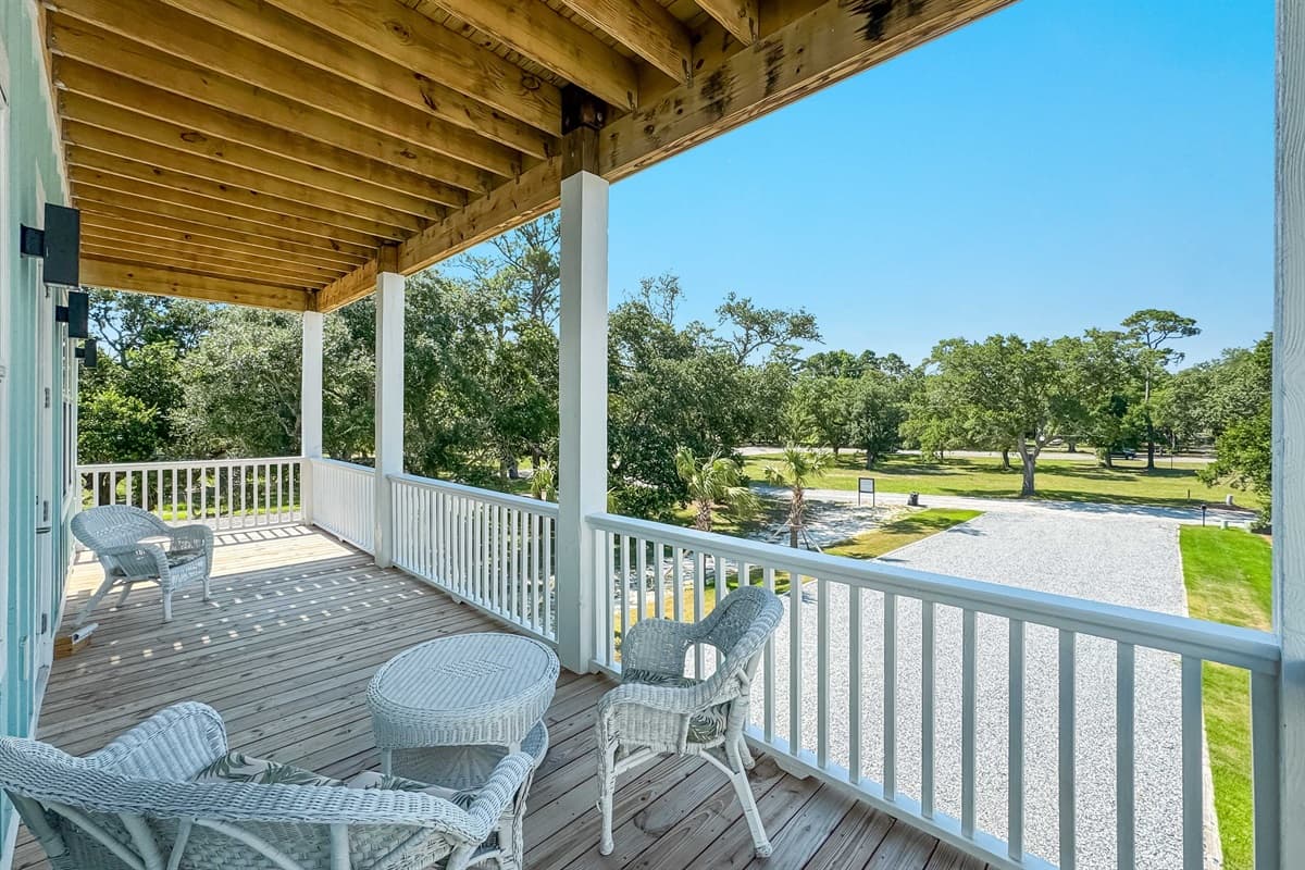 Enjoy fresh air and tranquility on this inviting covered deck, featuring comfortable wicker seating and a charming side table. Overlooking a large open area with trees, it's an ideal spot for relaxation and appreciating the peaceful environment.