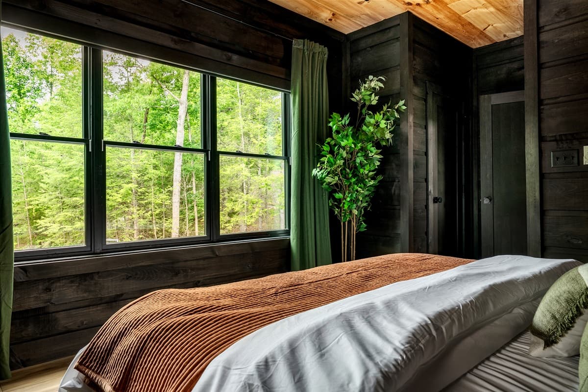 This inviting bedroom features large windows offering a lush green view, a comfortable bed, and a decorative plant adding to the serene ambiance.
