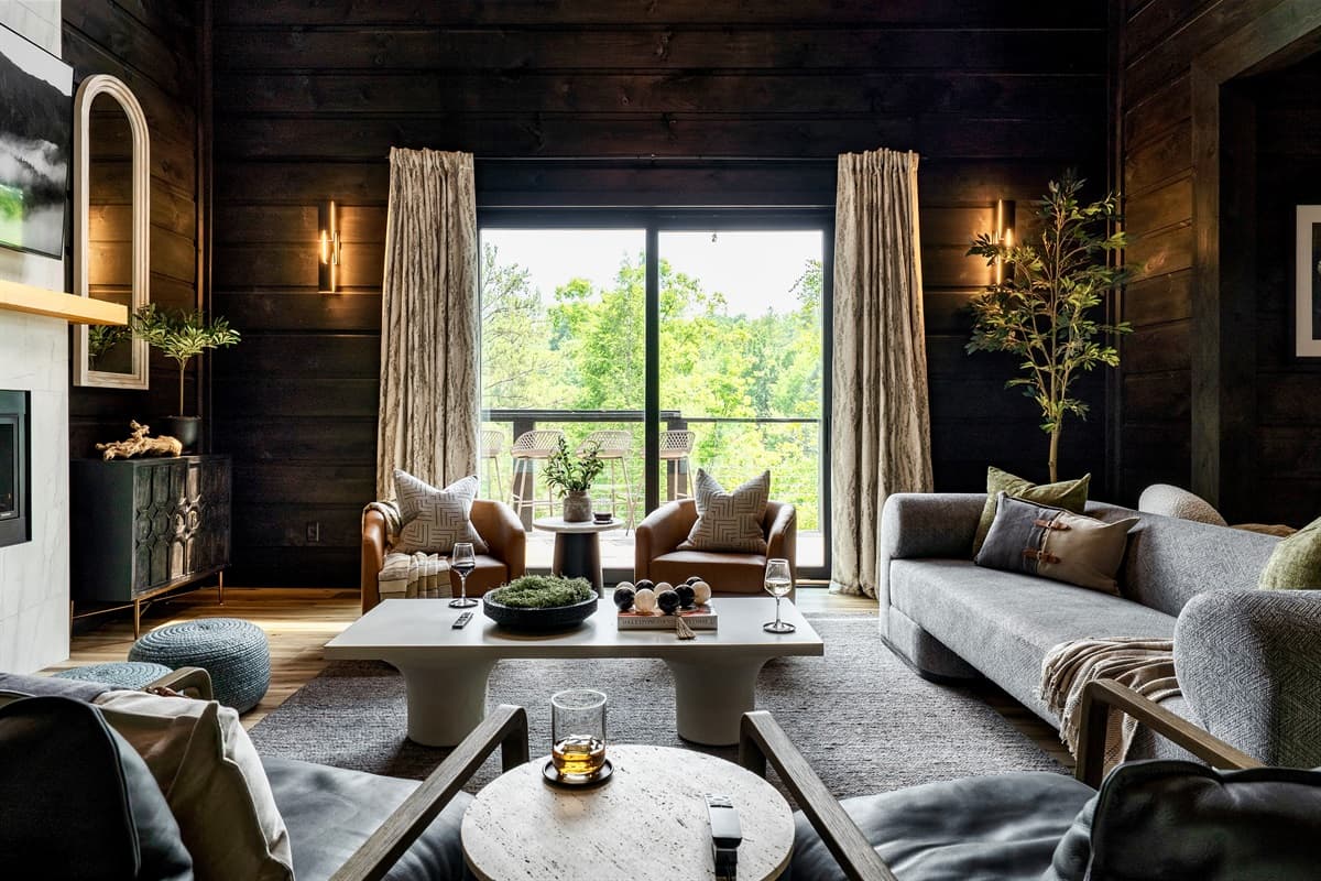 Relax and unwind in this beautifully appointed living space, showcasing a stylish grey sofa, chic armchairs, and a modern coffee table, all set against a backdrop of rich dark wood walls. Expansive windows frame picturesque views of the outdoors, creating