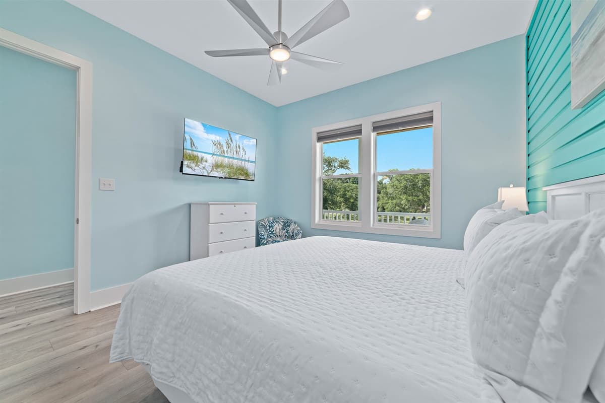 This bright bedroom features a comfortable queen-sized bed, a wall-mounted TV, and large windows with outdoor views.
