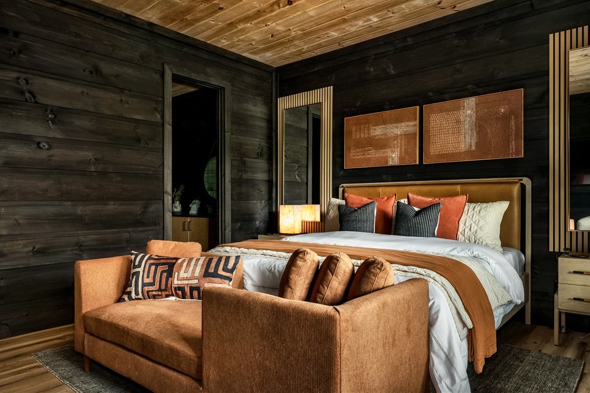 Relax in style in this beautifully designed bedroom, offering a plush bed, a cozy rust-colored sofa with patterned pillows, and a private ensuite bathroom accessible through a dark wooden door. The interplay of dark wooden walls, warm textures, and mirrore