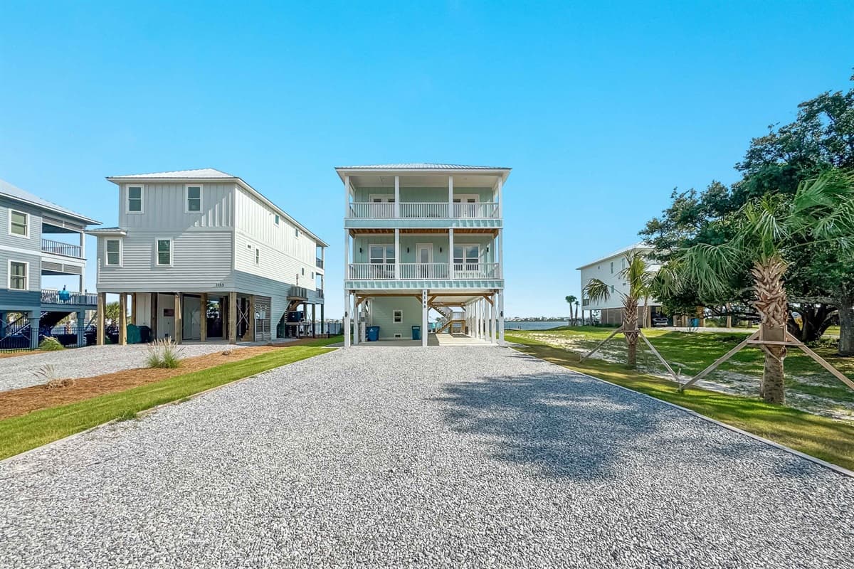 Welcome to this stunning property, showcasing a multi-story coastal residence with a vibrant blue-green exterior and charming wrap-around balconies. The large gravel driveway provides ample parking, while the surrounding natural landscape and distant bay v