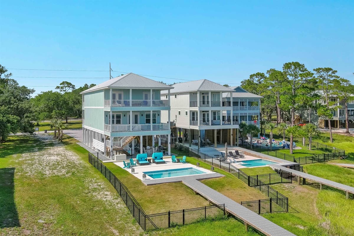 This stunning aerial view showcases two multi-level coastal homes, each with a private pool and spacious outdoor living areas, set against a backdrop of lush greenery and the bay.